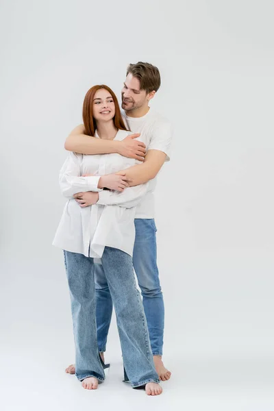 Усміхнений чоловік у джинсах, що обіймає позитивну дівчину в сорочці на білому тлі — стокове фото