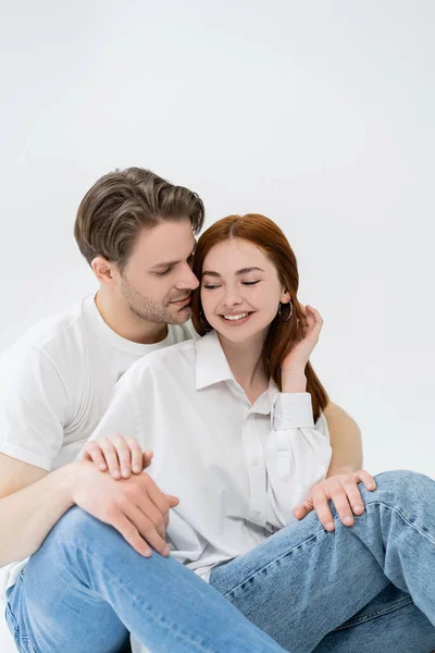 Junger Mann berührt glückliche Freundin in Hemd und Jeans auf weißem Hintergrund — Stockfoto