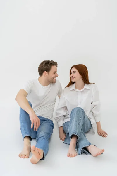 Pareja joven positiva en jeans mirándose mientras están sentados sobre fondo blanco - foto de stock