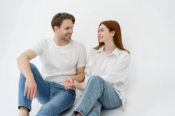 Coppia positiva in jeans che si tiene per mano mentre si siede su sfondo bianco — Foto stock