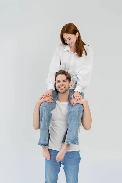 Cheerful man holding barefoot girlfriend on shoulders isolated on white — Stock Photo