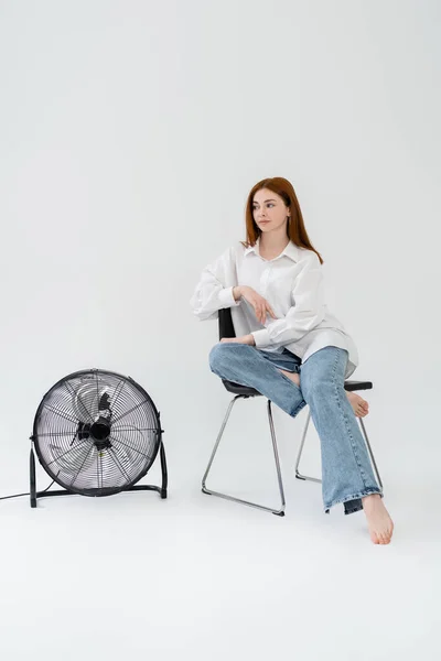 Jovem ruiva sentada na cadeira perto de ventilador elétrico no fundo branco — Fotografia de Stock