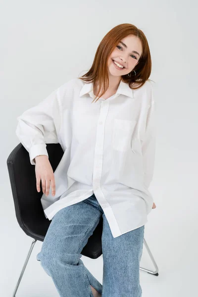 Fröhliche junge Frau im Hemd blickt in die Kamera, während sie auf einem Stuhl auf weißem Hintergrund sitzt — Stockfoto