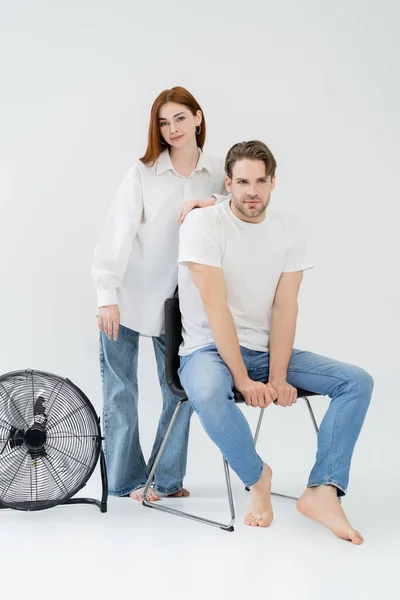 Jeune femme en chemise et jeans debout près du petit ami sur chaise sur fond blanc — Photo de stock