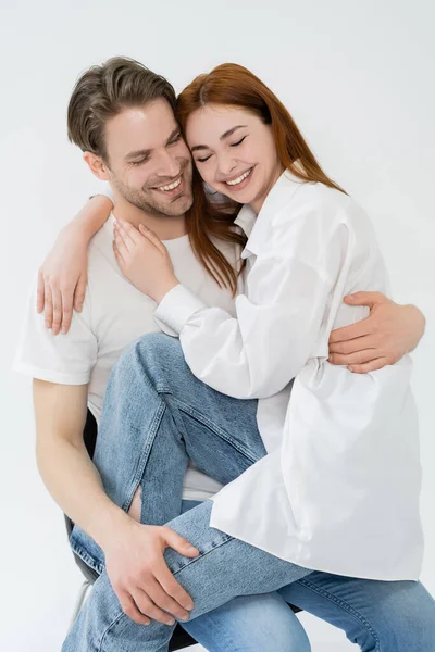 Pareja positiva abrazándose mientras está sentada en silla aislada en blanco - foto de stock