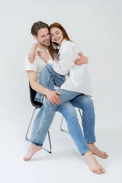 Glücklich barfuß Mann umarmt rothaarige Freundin, während sie auf Stuhl auf weißem Hintergrund sitzt — Stockfoto