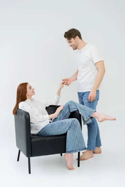 Side view of man holding hand of barefoot girlfriend sitting on armchair on white background — Stock Photo