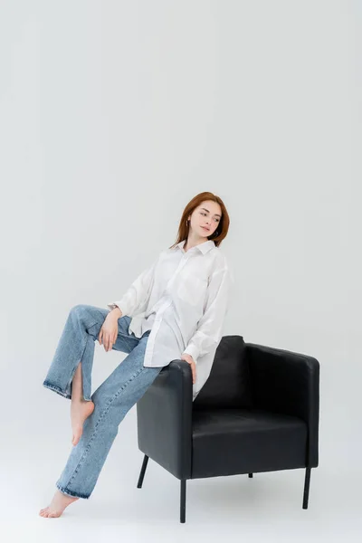 Femme pieds nus en jeans assis sur un fauteuil sur fond blanc — Photo de stock