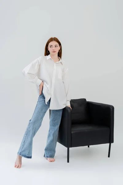 Pleine longueur de femme pieds nus en jeans et chemise debout près du fauteuil sur fond blanc — Photo de stock