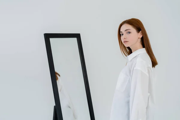 Молода руда волохата жінка в сорочці дивиться на камеру біля дзеркала ізольовано на білому — стокове фото