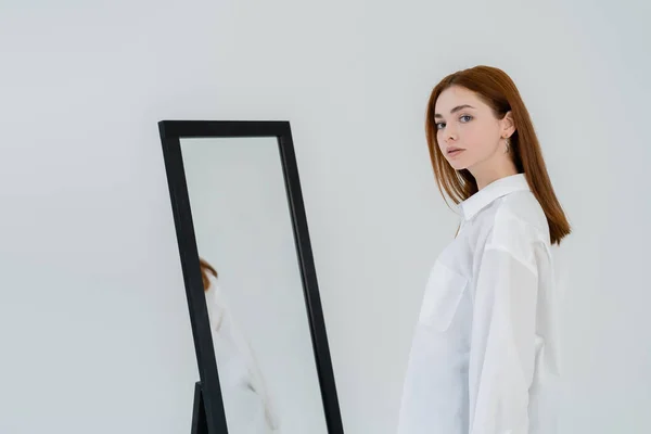 Jovem de cabelos vermelhos mulher na camisa olhando para a câmera perto do espelho isolado no branco — Fotografia de Stock