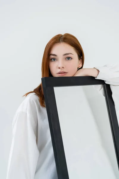 Руда жінка в сорочці дивиться на камеру біля дзеркала ізольовано на білому — стокове фото
