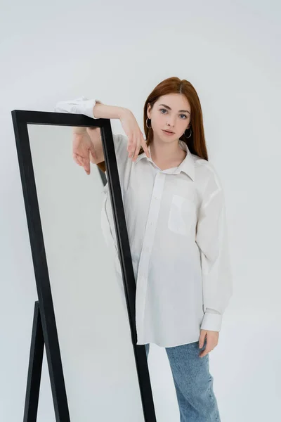 Mujer joven en camisa posteando cerca de espejo aislado en blanco - foto de stock