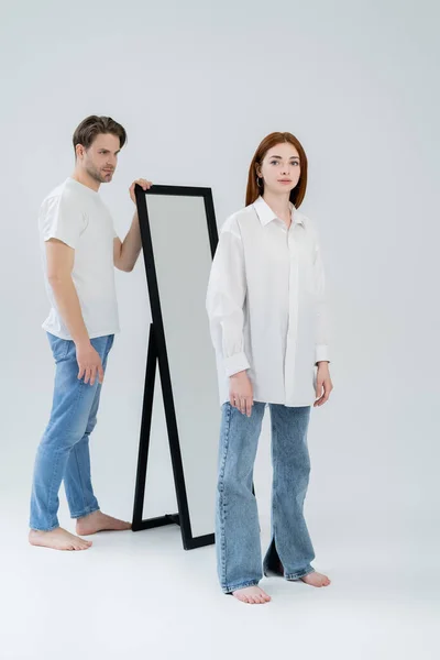 Pleine longueur de femme pieds nus regardant la caméra près du miroir et petit ami sur fond blanc — Photo de stock