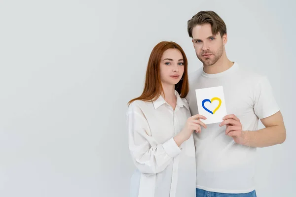 Jeune couple tenant la carte avec le signe de coeur bleu et jaune isolé sur blanc — Photo de stock