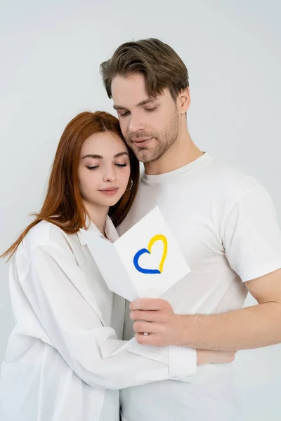 Homem segurando cartão com sinal de coração azul e amarelo perto da namorada isolada no branco — Fotografia de Stock