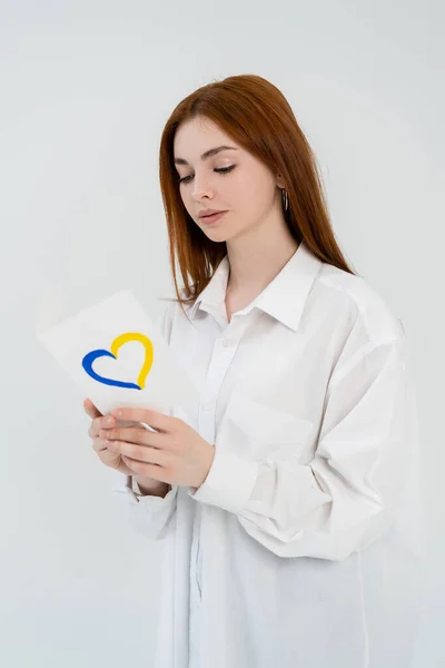 Femme rousse en chemise tenant la carte avec le signe de coeur bleu et jaune isolé sur blanc — Photo de stock
