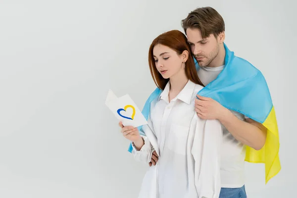 Hombre en bandera ucraniana abrazo novia con tarjeta corazón signo aislado en blanco - foto de stock