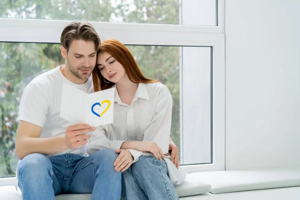 Man holding card with blue and yellow heart sign near girlfriend and window — Stock Photo