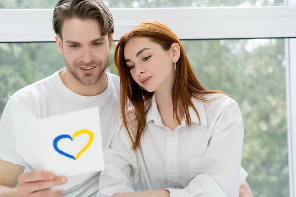 Homme souriant tenant la carte avec le signe de coeur bleu et jaune près de petite amie à la maison — Photo de stock