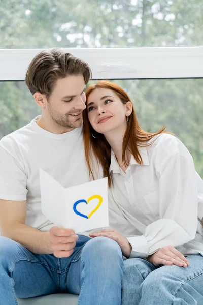 Positive man holding card with blue and yellow heart sign and looking at girlfriend at home — Stock Photo