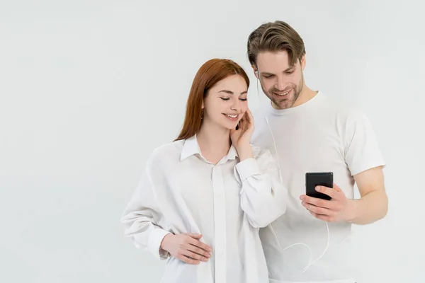 Pareja alegre usando teléfono celular y auriculares aislados en blanco - foto de stock