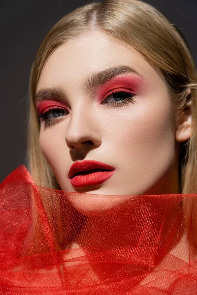 Donna con trucco rosso guardando macchina fotografica vicino tessuto di tulle isolato su grigio — Foto stock