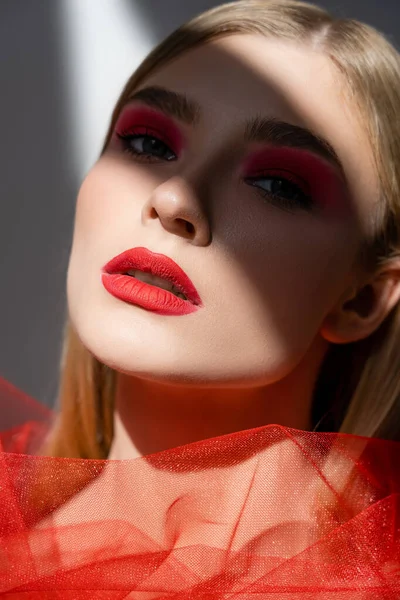 Young woman with red lips standing in shadow near tulle cloth on grey background — Stock Photo