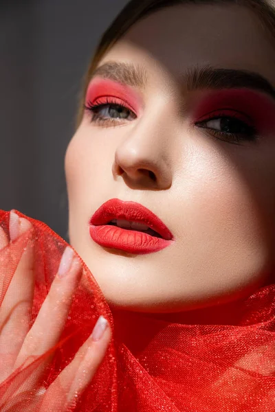 Mujer joven con labios rojos tocando tela de tul mientras está de pie en la sombra aislada en gris - foto de stock