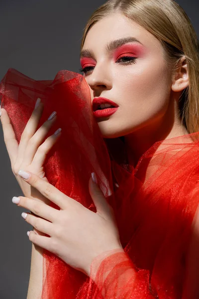 Mulher bonita com maquiagem segurando pano de tule vermelho isolado no cinza — Fotografia de Stock