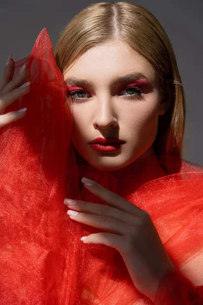 Retrato de mujer joven con maquillaje rojo sosteniendo un paño de tul y mirando a la cámara aislada en gris - foto de stock