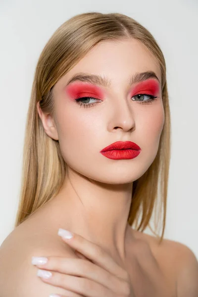 Mujer bonita con maquillaje rojo tocando hombro desnudo aislado en gris - foto de stock