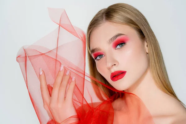 Mujer de pelo rubio con sombra de ojos rojos sosteniendo tela de tul aislada en gris - foto de stock