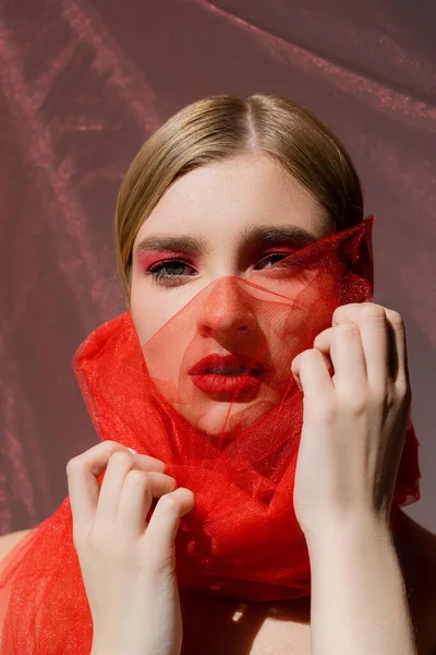 Modelo de pelo claro con sombra de ojos rojos tocando tela de tul sobre fondo gris - foto de stock