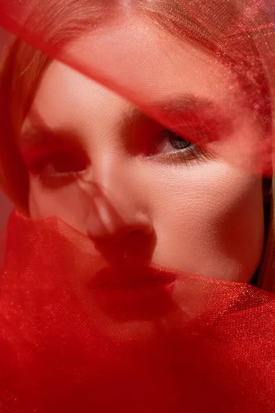 Close up view of young woman looking at camera near red blurred cloth — стоковое фото