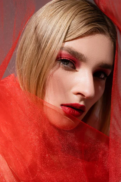 Modelo de pelo claro con sombra de ojos rojos posando cerca de tela sobre fondo gris - foto de stock