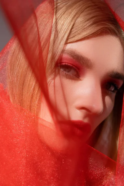 Modelo borroso con labios rojos mirando a la cámara cerca de tela sobre fondo gris - foto de stock