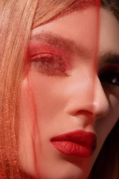 Close up view of woman with makeup looking at camera near blurred red cloth isolated on black — стоковое фото