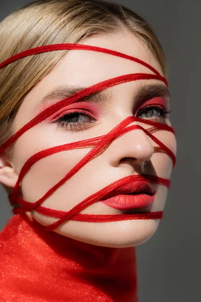 Portrait of woman with red eye shadow and threads on face looking at camera isolated on grey — стоковое фото