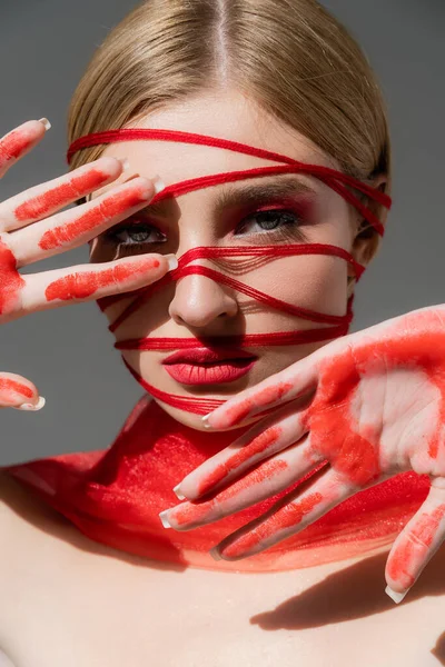 Jeune modèle avec des fils rouges sur le visage et de la peinture sur les mains en regardant la caméra isolée sur gris — Photo de stock