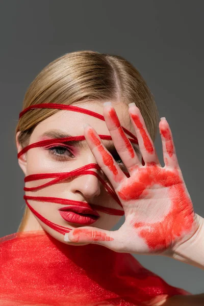 Светловолосая женщина с красной краской на руке и нитками на лице изолированы на сером — стоковое фото