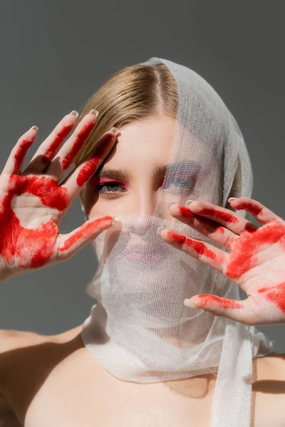 Mujer joven en vendaje médico en la cara y pintura en las manos mirando a la cámara aislada en gris - foto de stock