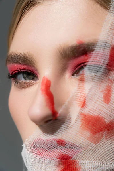 Vue rapprochée de la femme avec de la peinture rouge sur le bandage médical sur le visage regardant la caméra isolée sur le gris — Photo de stock