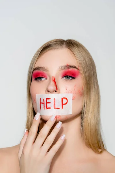 Mujer joven con sangre en la cara y tarjeta con letras de ayuda en los labios aislados en gris - foto de stock
