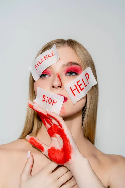 Jovem com visagem vermelha e papéis com silêncio, parar e ajudar a letras isoladas em cinza — Fotografia de Stock