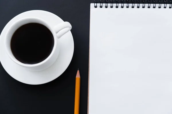 Top view of cup of coffee with saucer near notebook and pencil on black — Stock Photo