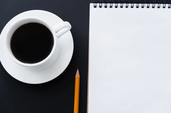 Top view of cup of coffee with saucer near notebook and pencil on black — стоковое фото