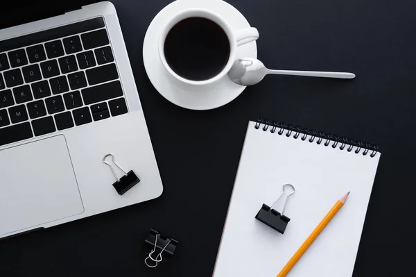 Top view of fold back clips and pencil on notebook near cup of coffee and laptop on black — стоковое фото