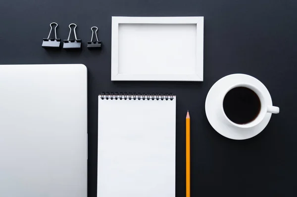 Flat lay of fold back clips near frame, blank notebook, laptop and cup of coffee on black — стоковое фото