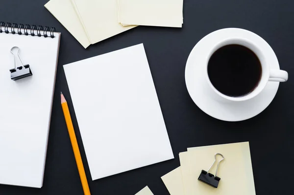 Top view of blank paper, notebook, pencil, fold back clips and cup of coffee on black — Stockfoto